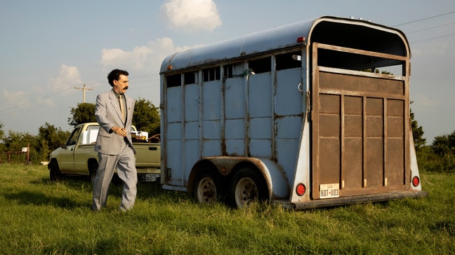 Borat standing beside a horse trailer in a grassy field