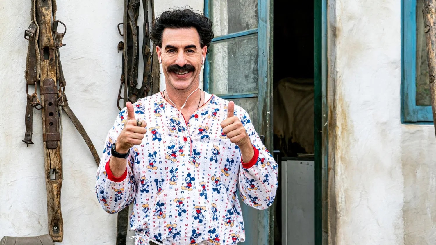 Borat standing outside a rustic building giving two thumbs up